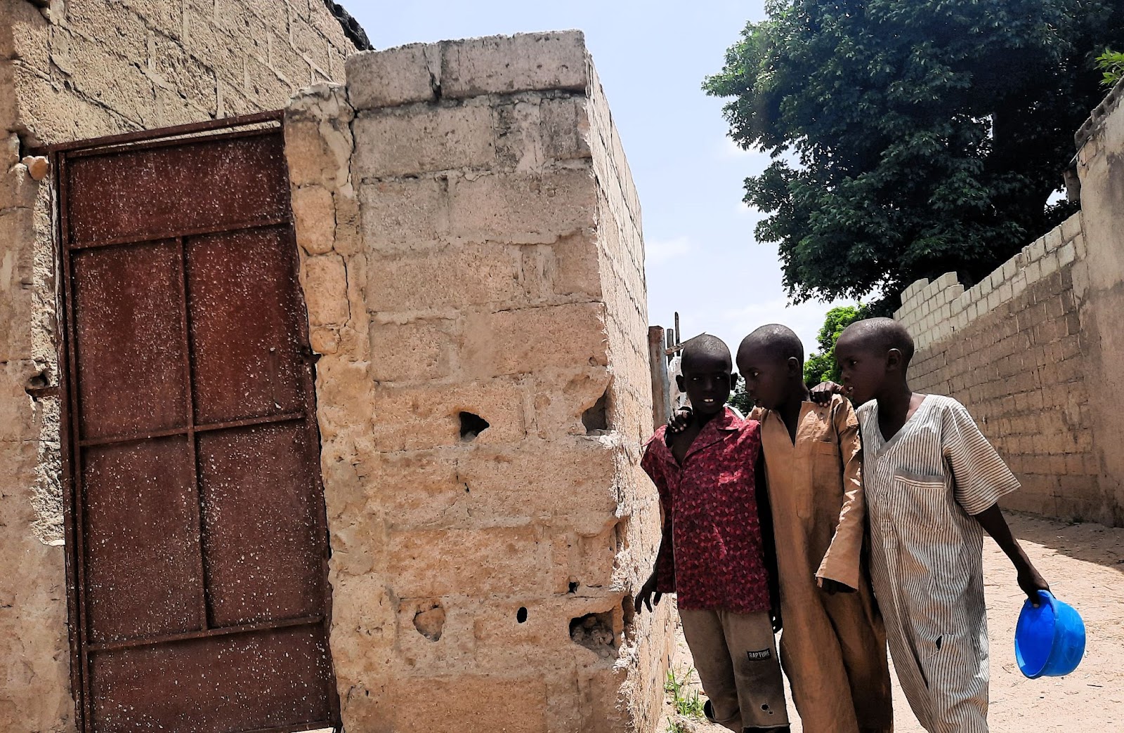 Three boys walking by a rusty door in a brick wall, one carrying a blue container.