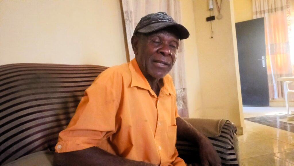 Elderly man in orange shirt and cap sitting on a couch, smiling casually indoors.