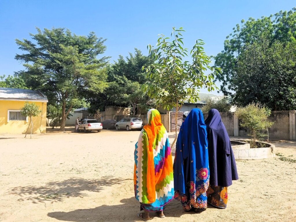 Three women in colorful traditional clothing standing in a sunny, sandy courtyard with trees and buildings in the background.