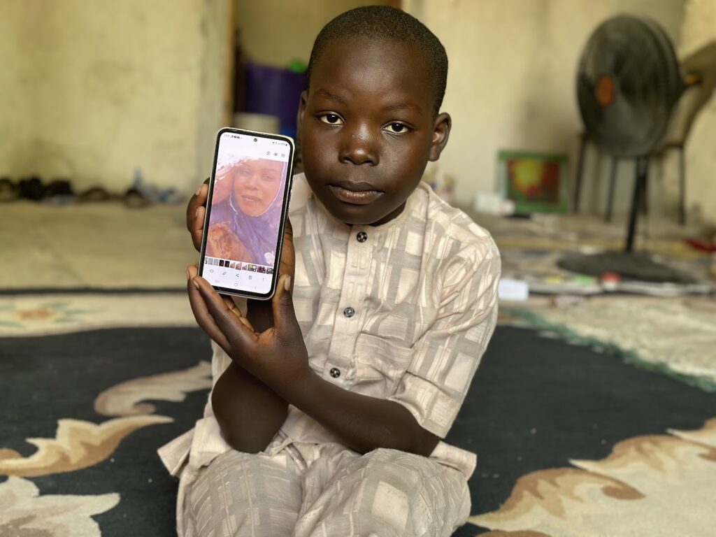 A young boy sitting indoors holds up a smartphone displaying a woman's photo on the screen.