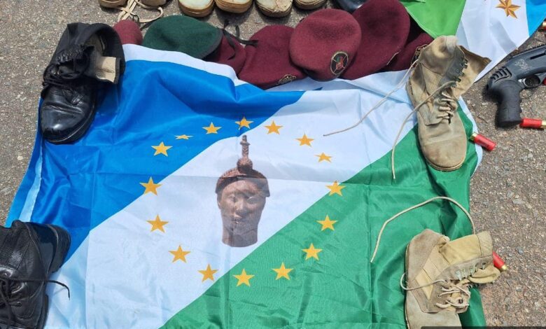 A flag with a central headpiece lying on the ground covered by various shoes, hats, and shotgun shells.