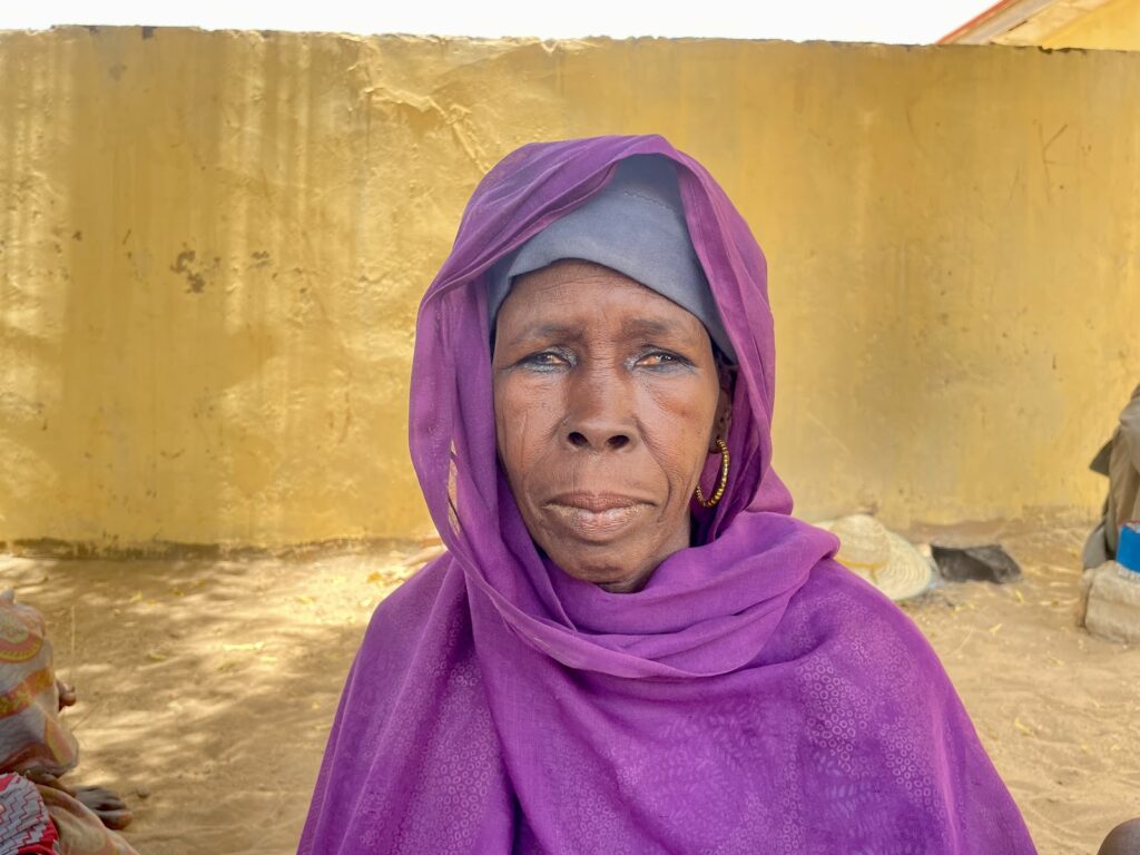Elderly woman in a purple headscarf with a contemplative expression against a yellow wall.