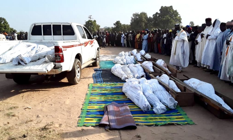 A group of people in traditional attire attending an outdoor ceremony with covered bodies laid out on the ground.