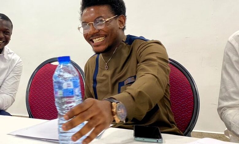 A smiling man with glasses reaching for a water bottle at a meeting, another person partially visible, with a phone and papers on the table.
