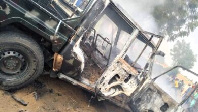 A burnt vehicle with open doors, smoke rising, and onlookers in the background.