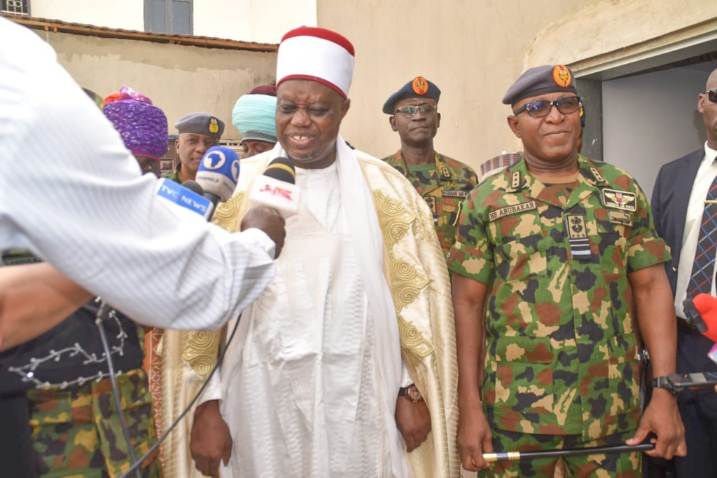 Man in traditional attire speaks to the press, flanked by military personnel.