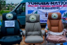 Three empty chairs in front of a van with a "Yoruba Nation" banner, symbolizing a protest or movement.