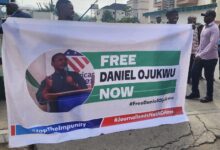 Banner held by people reading "FREE DANIEL OJUKWU NOW" with hashtags for activism and an image of a speaker.