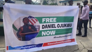 Banner held by people reading "FREE DANIEL OJUKWU NOW" with hashtags for activism and an image of a speaker.