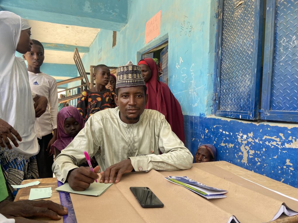 Man in traditional attire sits writing at a table in a classroom with children and adults looking on.