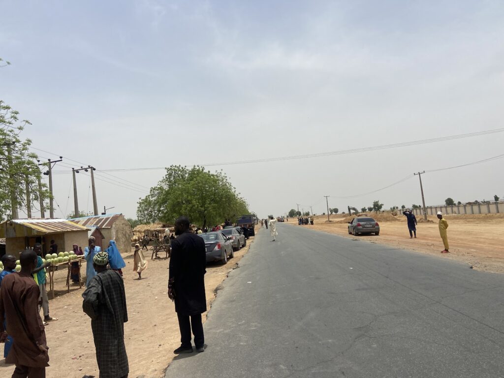 A busy roadside scene with people and vehicles in a semi-arid area under a hazy sky.