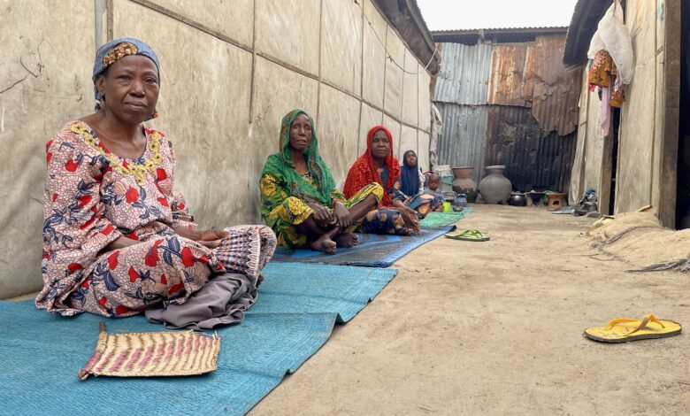 Women in traditional attire sitting outside on mats in a village alleyway.
