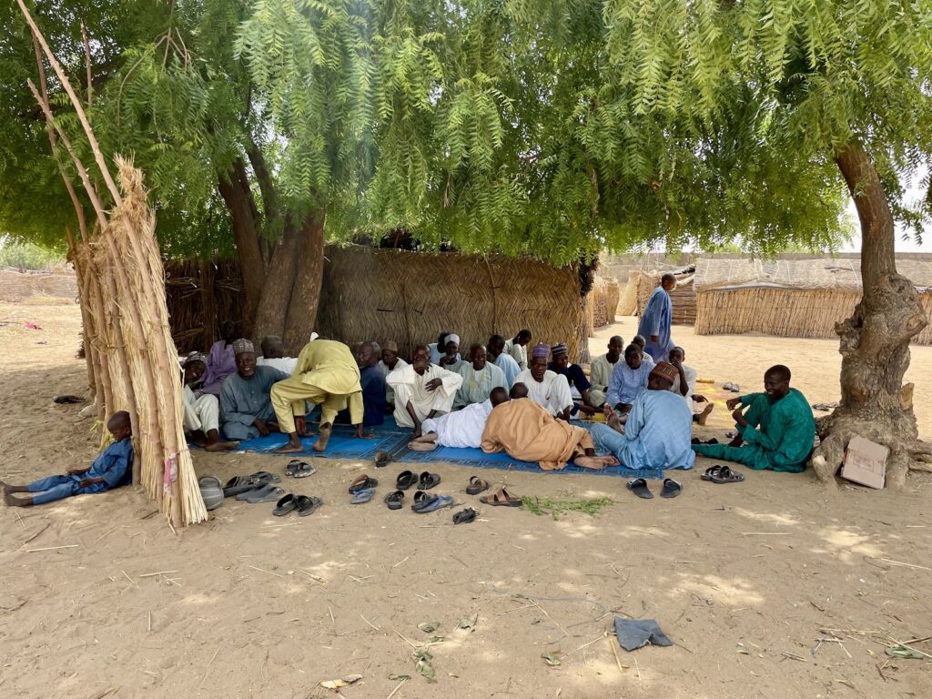 Group of men sitting under a tree in a rural setting, with shoes off and several engaged in conversation.