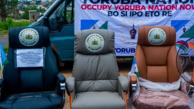 Three empty chairs in front of a van with a "Yoruba Nation" banner, symbolizing a protest or movement.