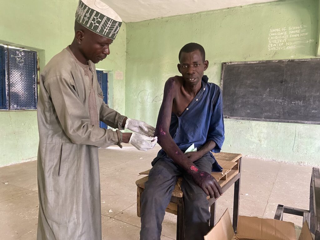 Man in traditional attire providing medical aid to another seated man with a bandaged arm in a classroom.