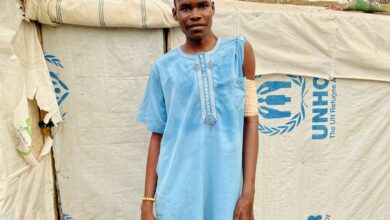 A young man in a blue garment stands before a tent with UNHCR logos.