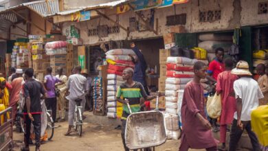 Bustling market scene with people shopping and transporting goods near stacked merchandise.