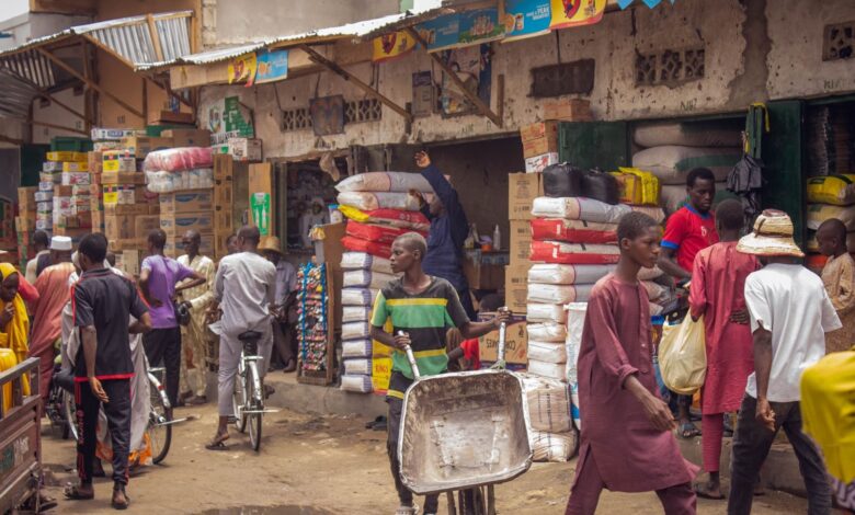 Bustling market scene with people shopping and transporting goods near stacked merchandise.