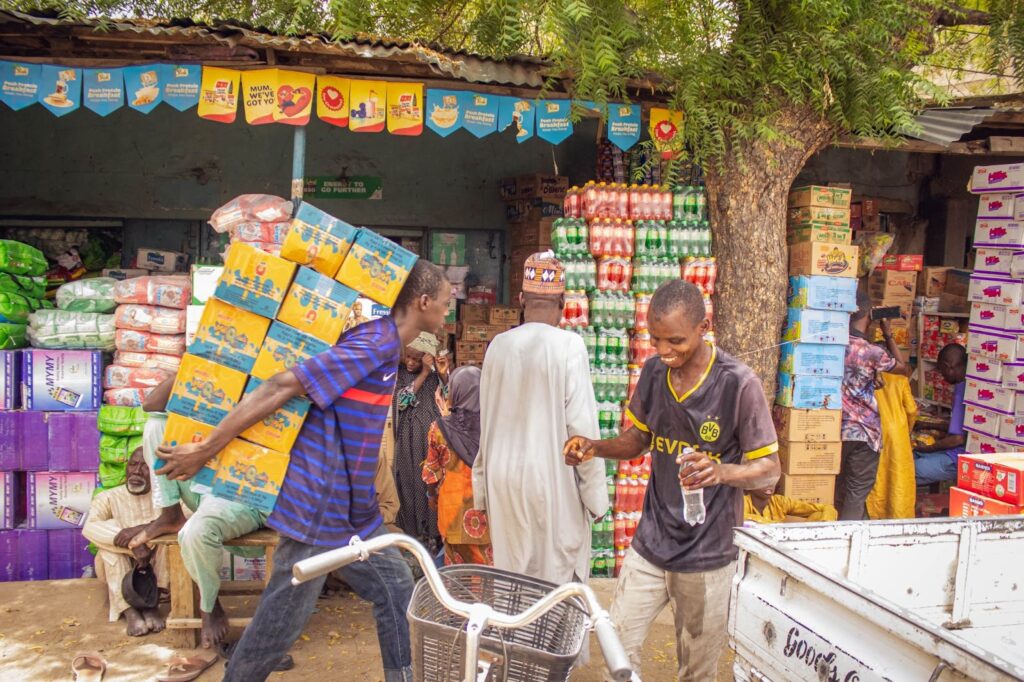 Vibrant outdoor market with people, products stacked high, and a man carrying a heavy load of boxed goods.