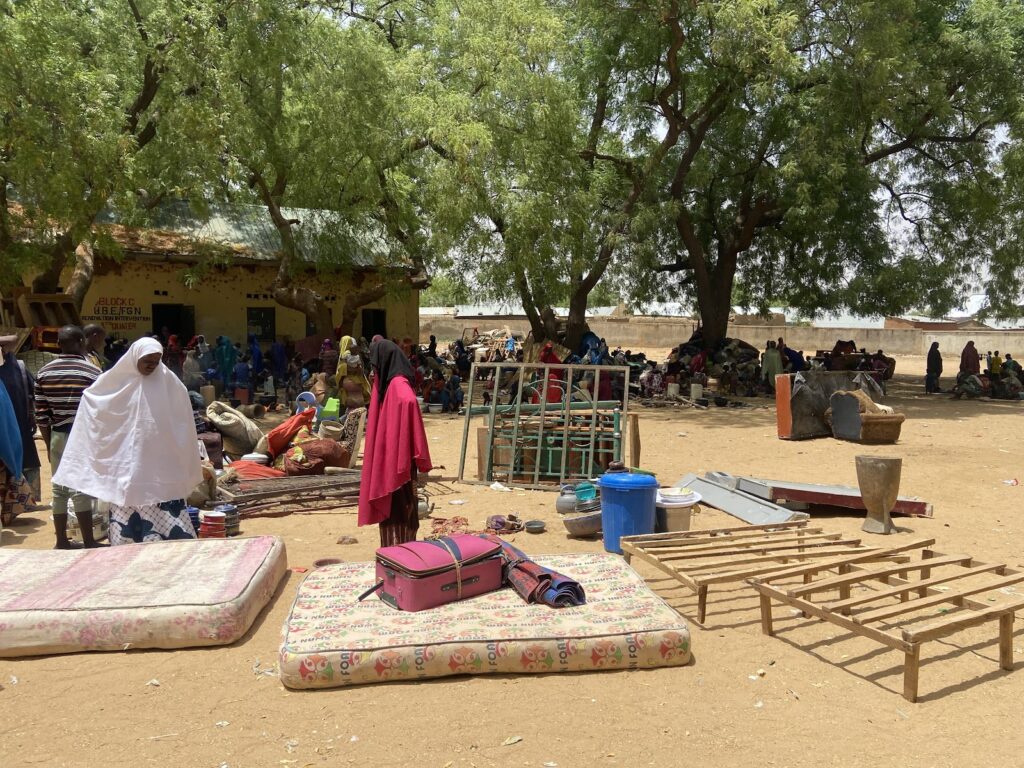 Outdoor market scene with people, mattresses, and various items under shady trees.