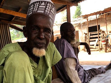 Elderly man in a patterned cap and green shirt with a pensive expression, another elder blurred in the background.