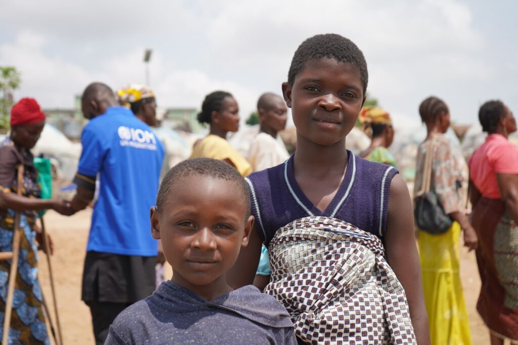 Two children smiling in a busy outdoor refugee camp setting.