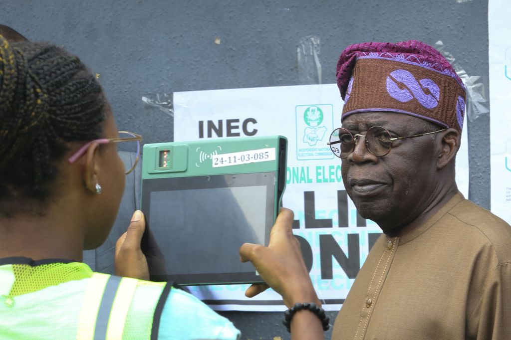 An election official uses a digital device to verify the identity of a man in traditional attire.