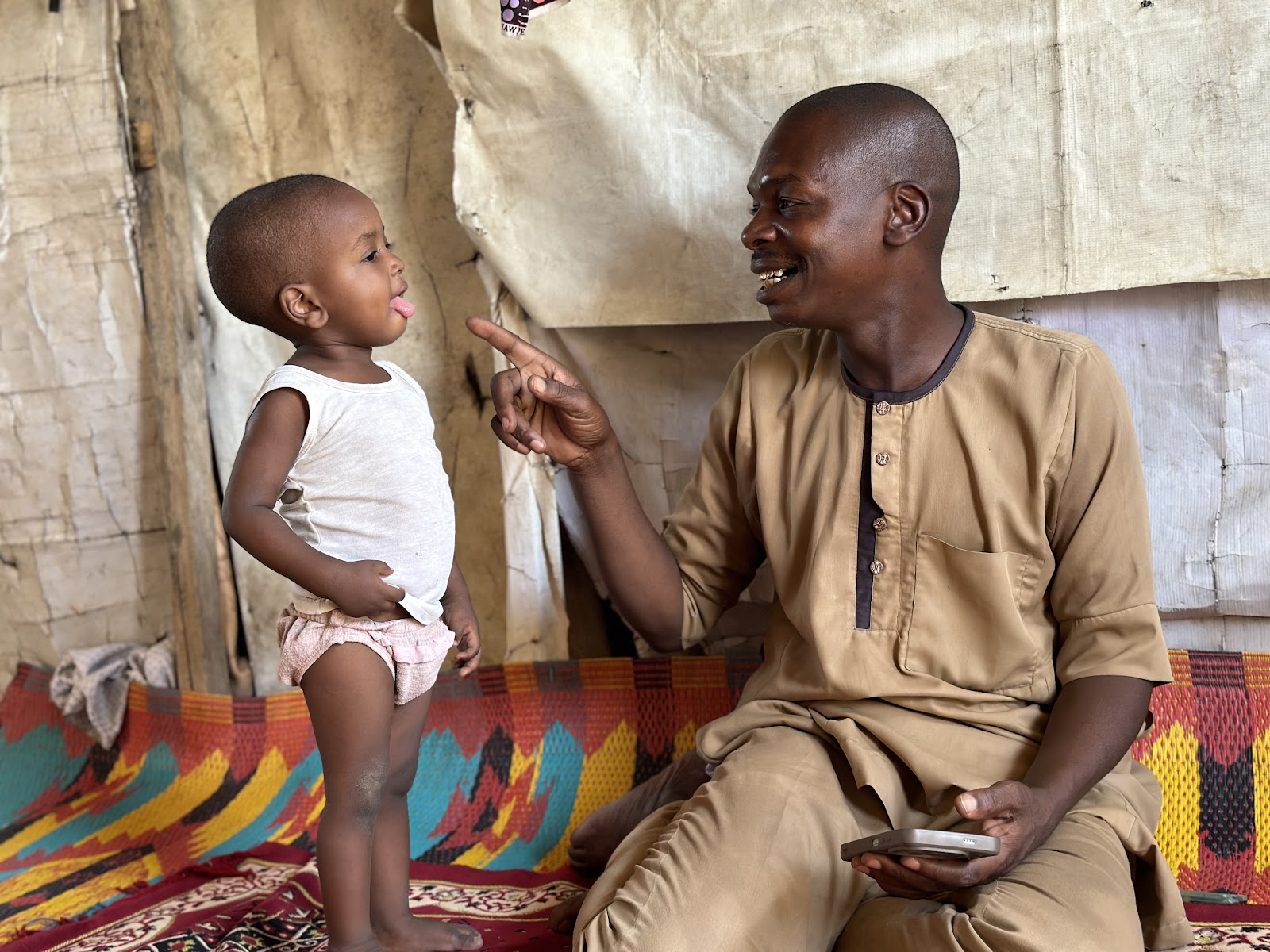 A joyful toddler sticking tongue out at a smiling man who is pointing at the child, sitting in a traditional dwelling.
