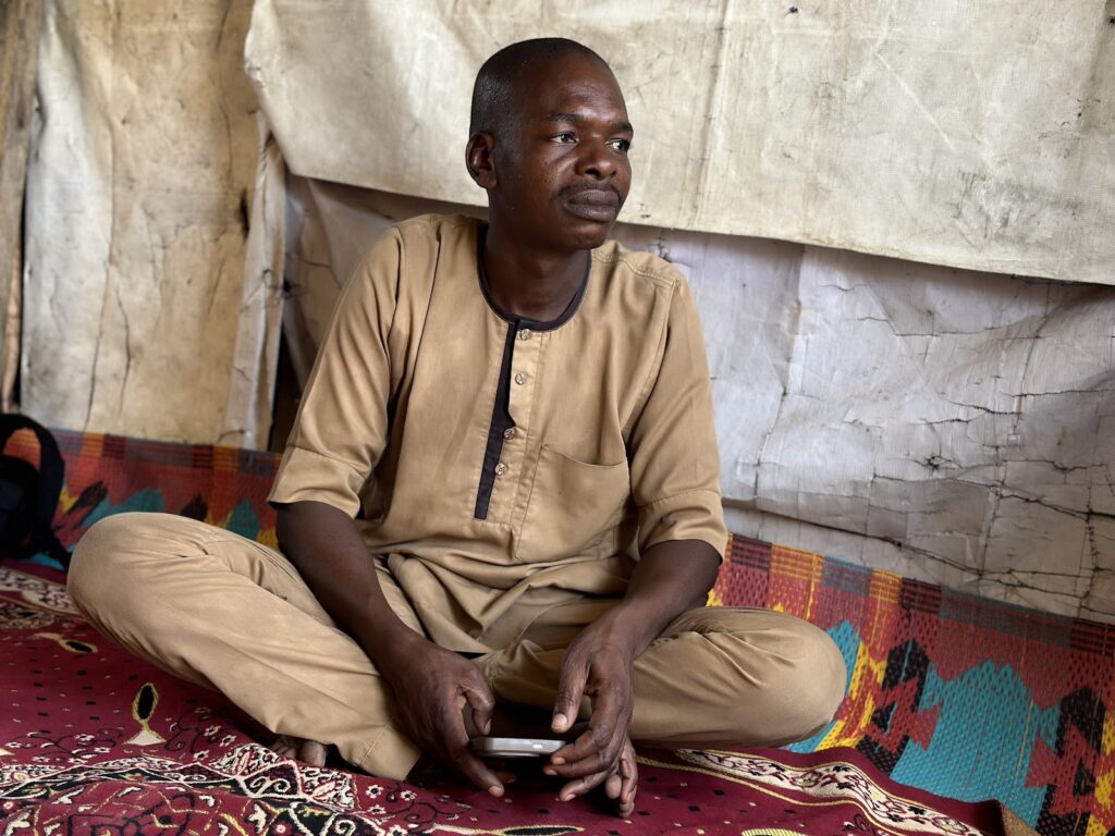 Man sitting cross-legged on a colorful rug, holding a phone, with a pensive expression, inside a tent.