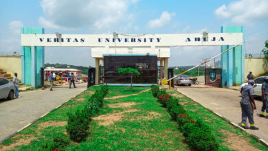 Entrance gate of Veritas University Abuja with people walking and cars parked nearby under a clear sky.