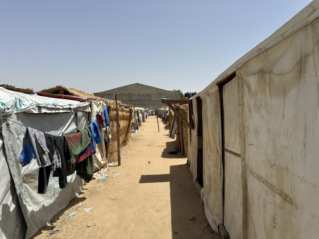A dusty pathway between rows of makeshift tents with laundry hanging outside under a clear sky.