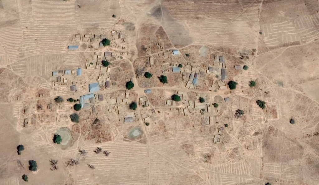 Aerial view of a sparse village with small buildings and trees amid dry, patterned fields.