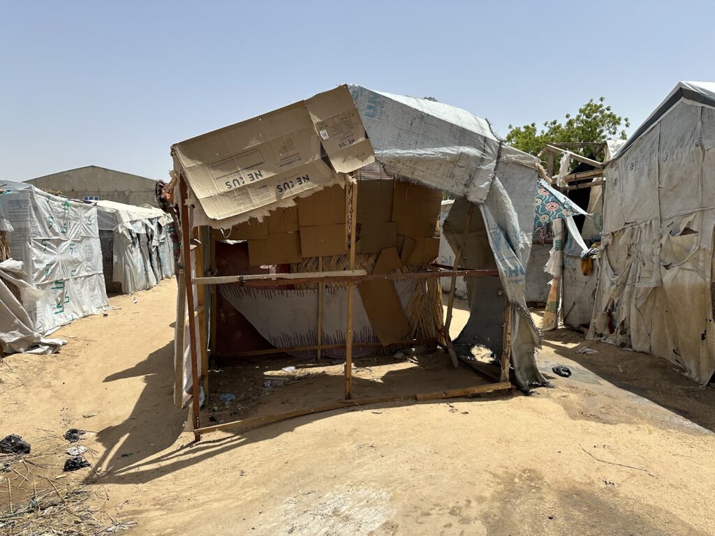 Makeshift shelters made of cardboard and tarp in a dusty environment suggesting an informal settlement or refugee camp.