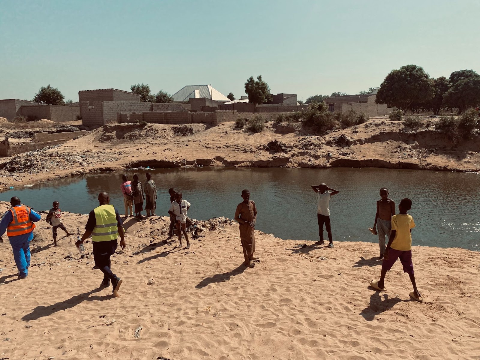 People gathered by a small river with sandy banks, buildings and trees in the background.