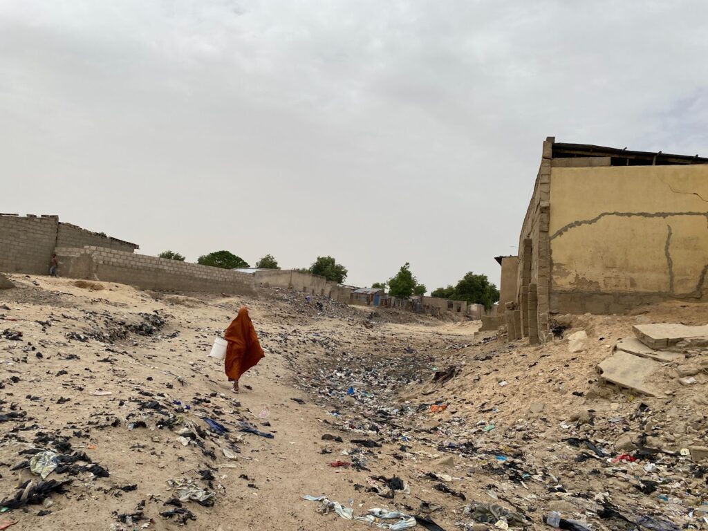 Person in orange clothing walking through a littered field near dilapidated buildings under a cloudy sky.