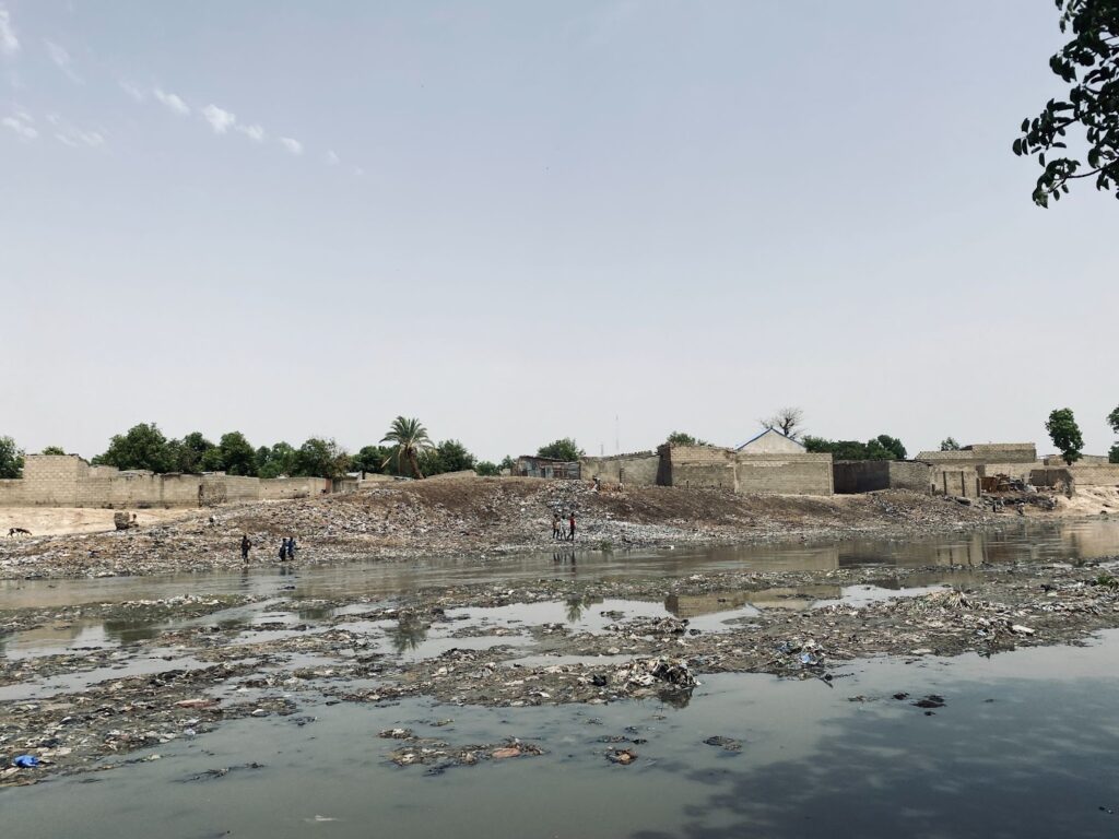 Polluted water body with litter, people walking nearby, and buildings at the backdrop under a clear sky.