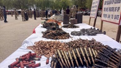 Table with seized ammunition and weapons on display, military personnel in background, outdoors under daylight.