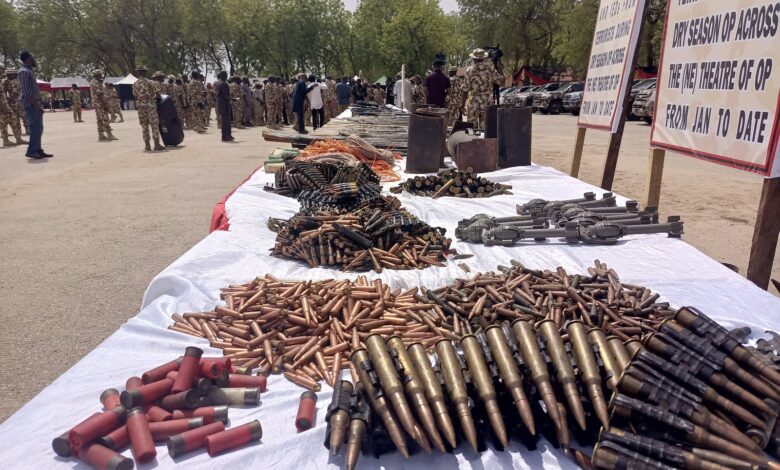 Table with seized ammunition and weapons on display, military personnel in background, outdoors under daylight.