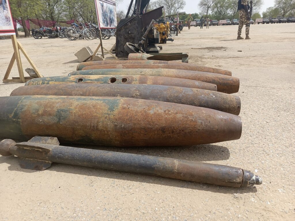 Rusty artillery shells lined up on ground with a military vehicle and motorcycles in the background.