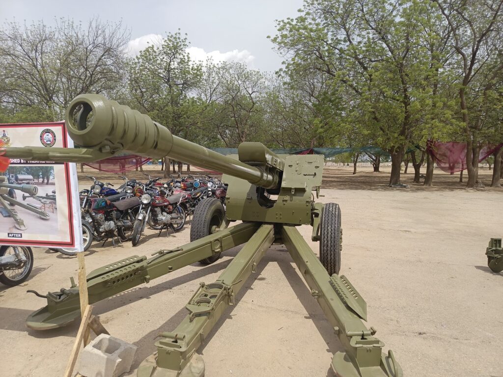 A green artillery cannon on display outdoors with motorcycles and trees in the background.