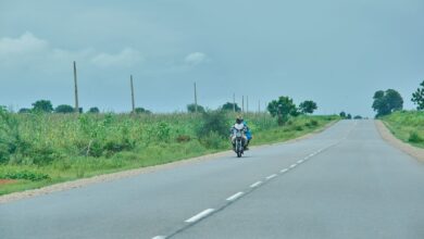 Motorcyclist riding on a rural road with greenery on both sides under a cloudy sky.