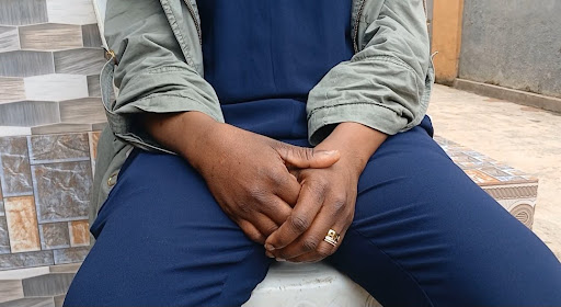 Close-up of a seated person's clasped hands on their lap, wearing blue trousers and a green jacket.