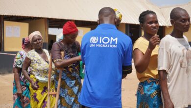 IOM staff member in blue shirt with locals in a sunny outdoor setting, likely during a migration-related activity.