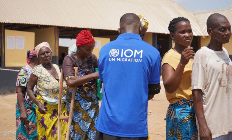 IOM staff member in blue shirt with locals in a sunny outdoor setting, likely during a migration-related activity.