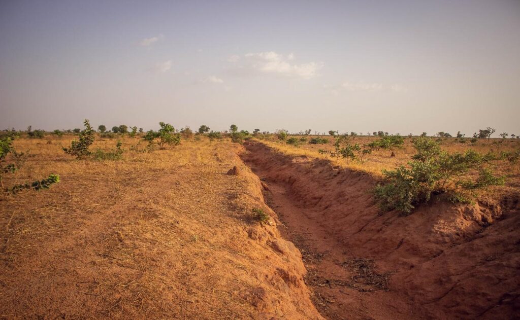 Dry landscape with a deep erosion gully cutting through arid soil and sparse vegetation under a hazy sky.