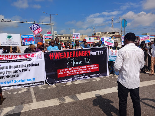 Group of protesters holding banners and signs at a rally with a person viewed from behind taking a photo.