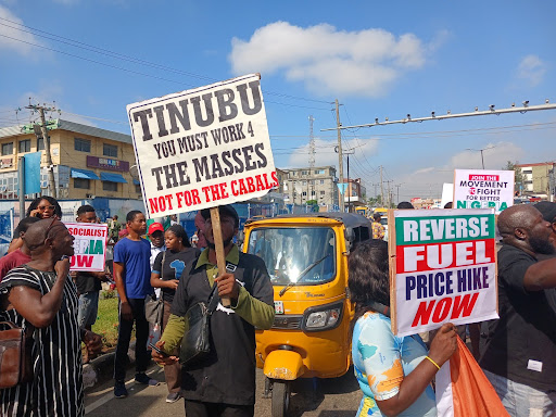 Protesters holding signs demanding political change and reversal of fuel price hike on a busy street.