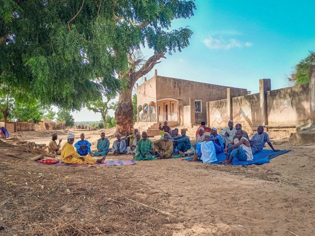 A group of people sitting under a tree near a traditional building in a sandy area, appearing to be in conversation.