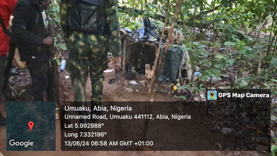 Two individuals in camouflage near a makeshift camp in the woods, with geo-location watermark for Umuaku, Nigeria.