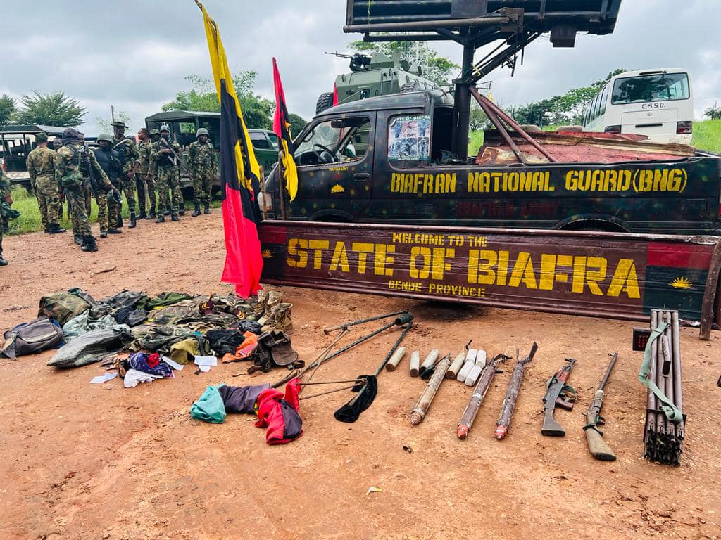 Military personnel stand by a vehicle with "Biafran National Guard" text, flanked by flags, with assorted gear and weapons on the ground.